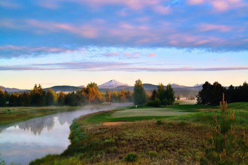 A serene golf course at sunrise, with mist over a river and mountains in the background under a colorful sky.