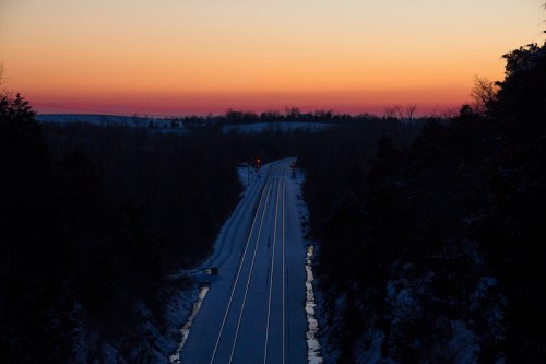 A serene winter landscape at dusk, featuring snow-covered train tracks leading into a colorful sunset.