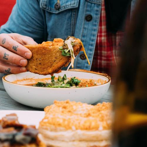 A person holds a sandwich over a bowl of soup, with a plate of food in the foreground.