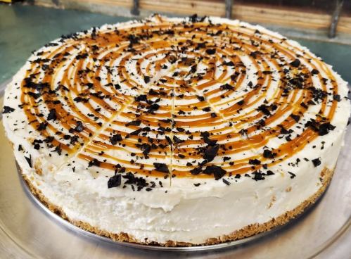 A round cheesecake topped with caramel swirls and dark chocolate flakes on a silver platter.