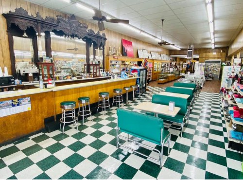 A vintage diner interior with a checkered floor, counter stools, and green booths, featuring wooden decor and display cases.