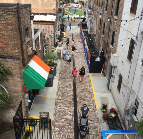 A cobblestone alley with people walking, shops, and string lights, featuring an Irish flag on a nearby building.