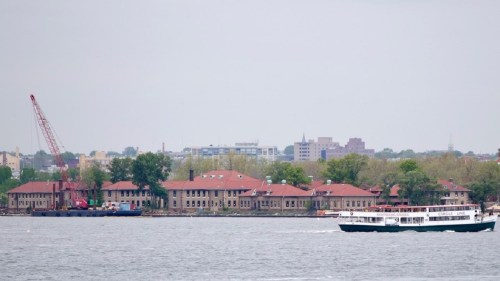 A ferry sails past a waterfront with buildings and a crane in the background under a cloudy sky.