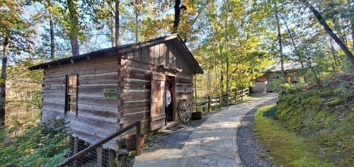 A rustic wooden cabin beside a stone path, surrounded by trees and greenery in a serene outdoor setting.