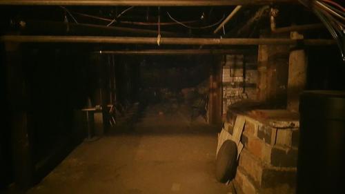 Dimly lit basement with exposed pipes, scattered debris, and a concrete floor. Shadows create an eerie atmosphere.