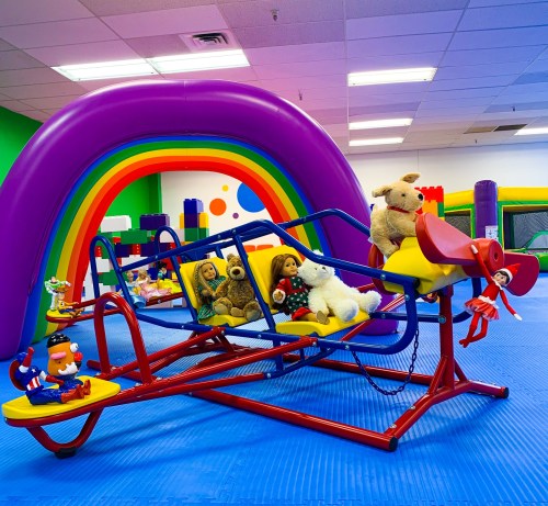 A colorful indoor play area featuring a playground set with stuffed animals and a rainbow arch in the background.