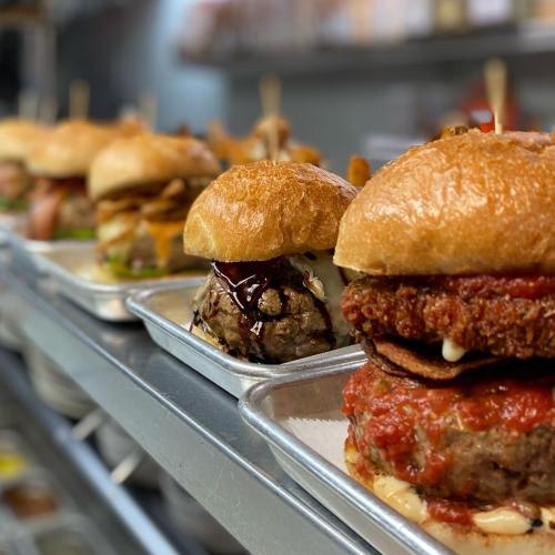 A row of gourmet burgers on metal trays, featuring various toppings and sauces, in a restaurant kitchen setting.