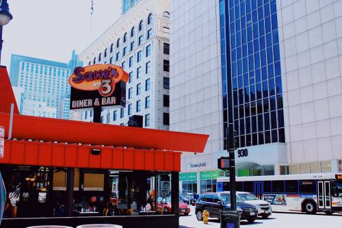 A diner with a red awning and sign reading "Sam's Diner & Bar" next to modern buildings and busy street traffic.