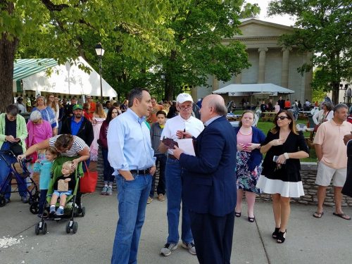 A group of people at an outdoor event, with two men engaged in conversation and others socializing in the background.