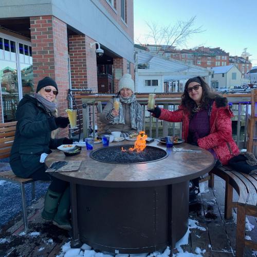 Three women sitting at a fire pit table outdoors, enjoying drinks and food in a winter setting.