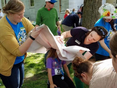 Two women assist a girl sitting on the ground, wrapping her head with paper during an outdoor event.