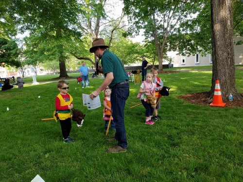 A group of children in costumes participate in an outdoor activity, guided by an adult in a hat. Trees and a building are in the background.