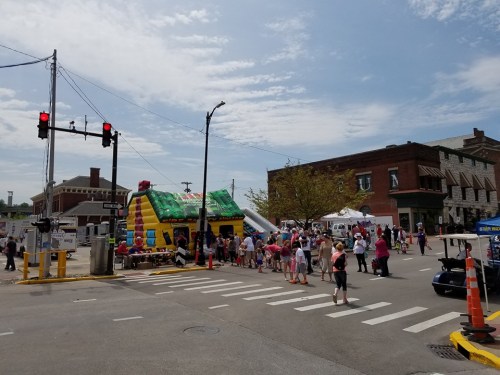 A lively street scene with a bounce house, children playing, and vendors at a community event on a sunny day.