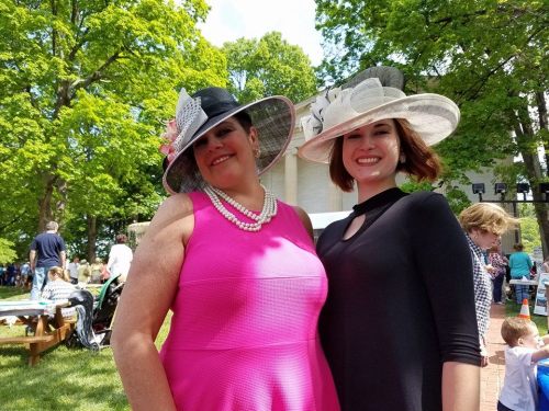 Two women in stylish hats and dresses pose together outdoors, surrounded by greenery and a lively crowd.