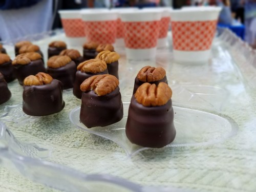 A tray of chocolate-covered treats topped with pecans, with paper cups in the background.