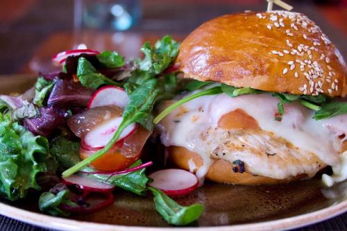 A sesame seed bun sandwich with grilled chicken, melted cheese, and a side salad of mixed greens and radishes.