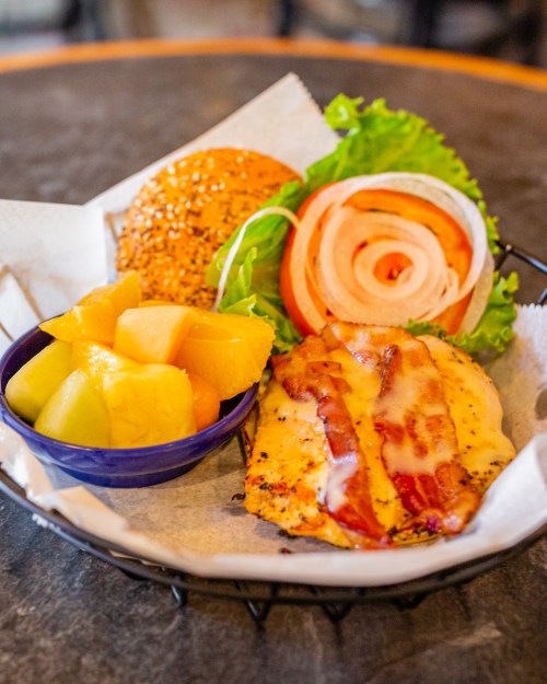 A plate featuring a grilled chicken sandwich with bacon, lettuce, tomato, and a side of mixed fruit.