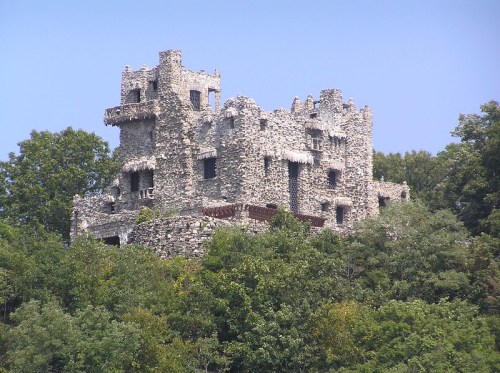 A stone castle-like structure perched on a hillside, surrounded by lush green trees under a clear blue sky.