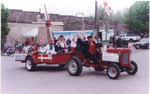A decorated tractor in a parade, featuring a windmill and people in traditional attire, celebrating Danish heritage.