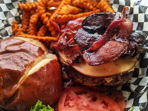 A juicy burger topped with bacon and cheese, served with curly fries, lettuce, and tomato on a checkered paper.