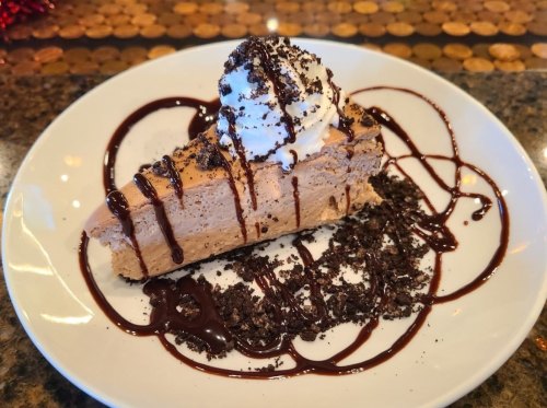 A slice of creamy dessert topped with whipped cream, chocolate drizzle, and crushed cookies on a white plate.