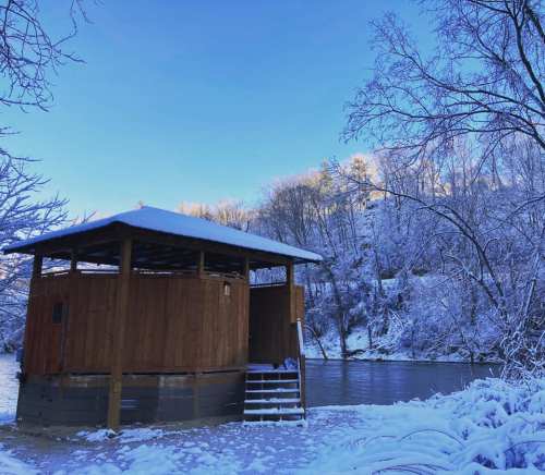 A wooden structure by a snowy river, surrounded by trees blanketed in white under a clear blue sky.