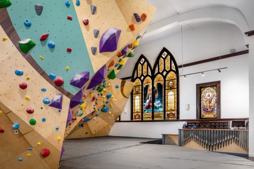 A colorful indoor climbing wall in a church, featuring stained glass windows and a wooden floor.