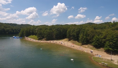 A serene lakeside scene with sandy beach, people relaxing, and lush green trees under a blue sky with fluffy clouds.