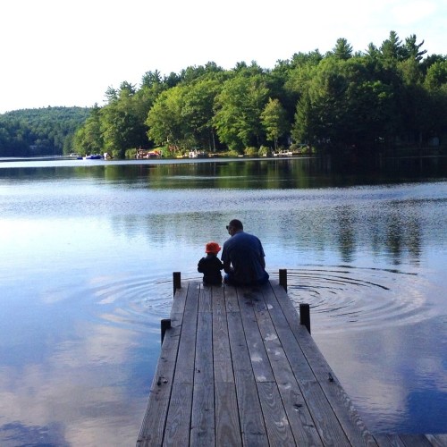 A person and a child sit on a wooden dock by a calm lake, surrounded by trees and a serene landscape.