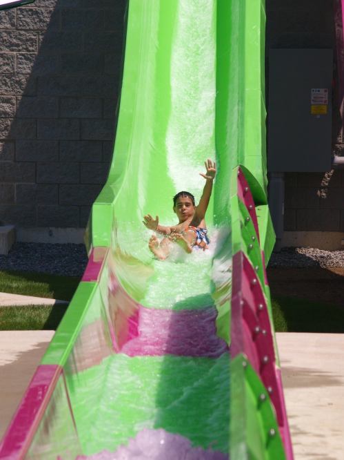 A child slides down a bright green water slide, splashing into a pool of water below.