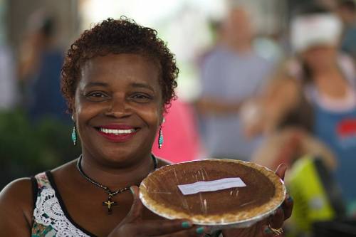 A smiling woman holds a pie, showcasing it proudly in a vibrant outdoor market setting.