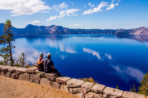 A couple sits on a stone wall, gazing at the serene blue waters of Crater Lake surrounded by mountains and trees.