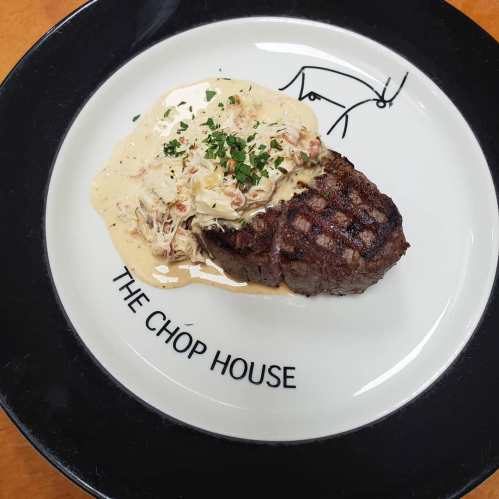 A grilled steak topped with creamy sauce and herbs, served on a plate with "The Chop House" logo.