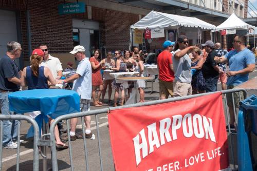 A lively outdoor event with people socializing near tables, a tent, and a prominent Harpoon banner in the foreground.