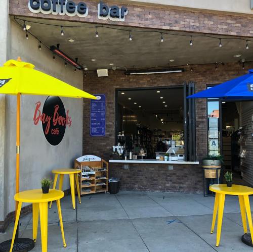 A coffee bar entrance with yellow and blue umbrellas, small tables, and a sign for Bay Books café.