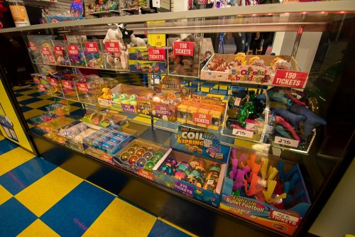 A display case filled with colorful toys and prizes, labeled with ticket values, in a vibrant arcade setting.
