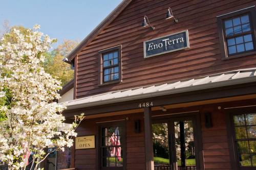 A rustic brown building with a sign reading "Eno Terra," surrounded by blooming flowers and trees.
