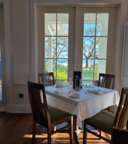 A dining table set for four, with a view of trees and a beach through large windows.