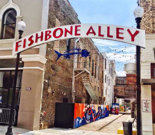 Sign for Fishbone Alley, a vibrant alleyway with colorful murals and string lights, surrounded by brick buildings.