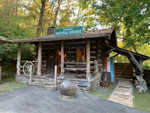 A rustic log cabin with a sign reading "Natural Bridge," surrounded by trees and a stone pathway.