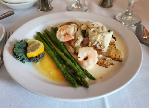 A plate of fish topped with crab and shrimp, served with asparagus and a lemon wedge on a white tablecloth.