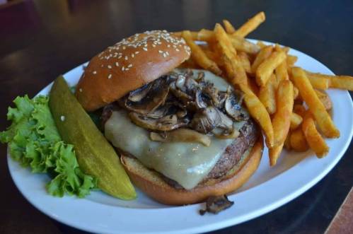 A juicy burger with melted cheese and sautéed mushrooms, served with fries, lettuce, and a pickle on a white plate.