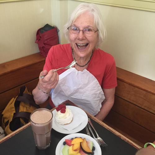 An elderly woman smiles while enjoying dessert and a drink at a café, wearing a bib and holding a fork.