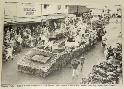 A decorated float passes by festival-goers, with a general store and vehicles in the background.