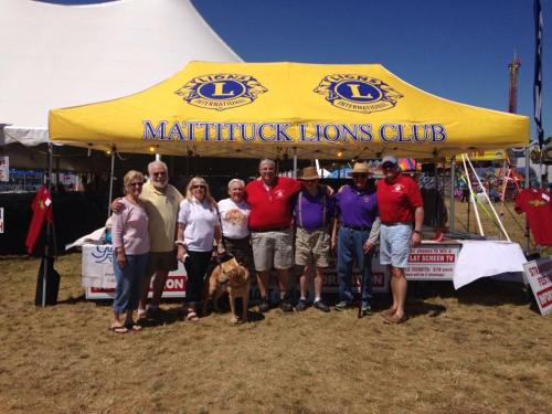 A group of people stands in front of a tent for the Mattituck Lions Club at an outdoor event, with a dog nearby.