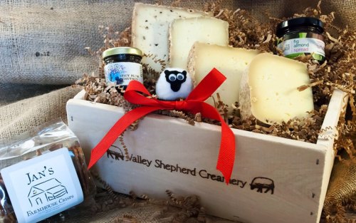 A wooden gift box filled with assorted cheeses, spreads, and a decorative sheep, all nestled in shredded paper.