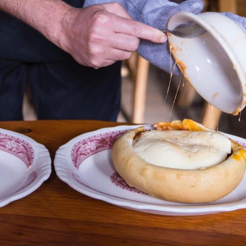 A person serves a cheesy dish from a bowl onto a plate, with melted cheese stretching as it's poured.