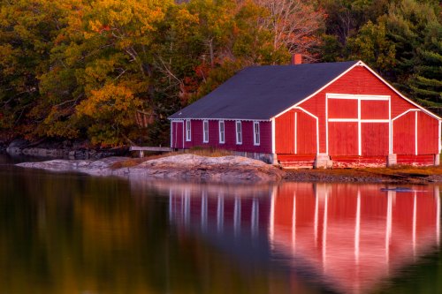 A red barn by a calm lake, surrounded by colorful autumn trees, reflecting in the water at sunset.
