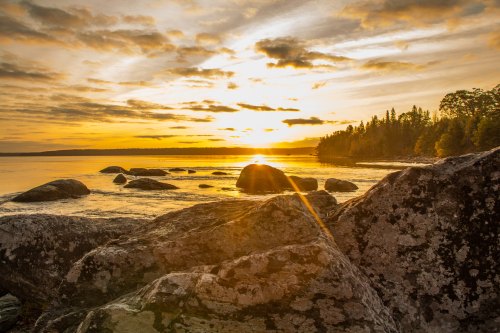 Sunset over a tranquil lake, with rocks in the foreground and trees lining the shore under a colorful sky.