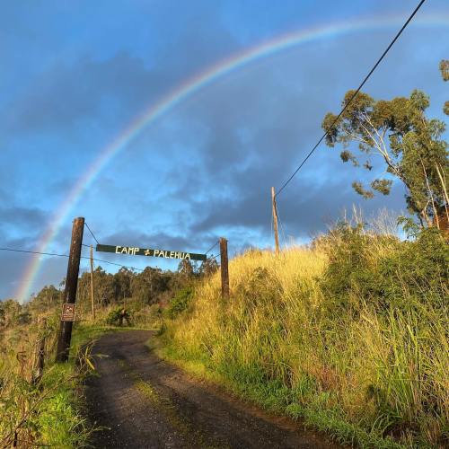 A scenic view of a road leading to "Camp Palehua" under a vibrant rainbow and blue sky, surrounded by lush greenery.